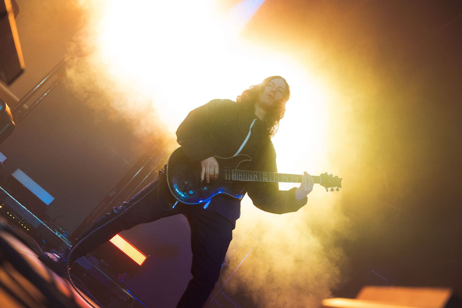 A guitarist performs on stage with dramatic backlighting creating a silhouette against a bright golden glow.