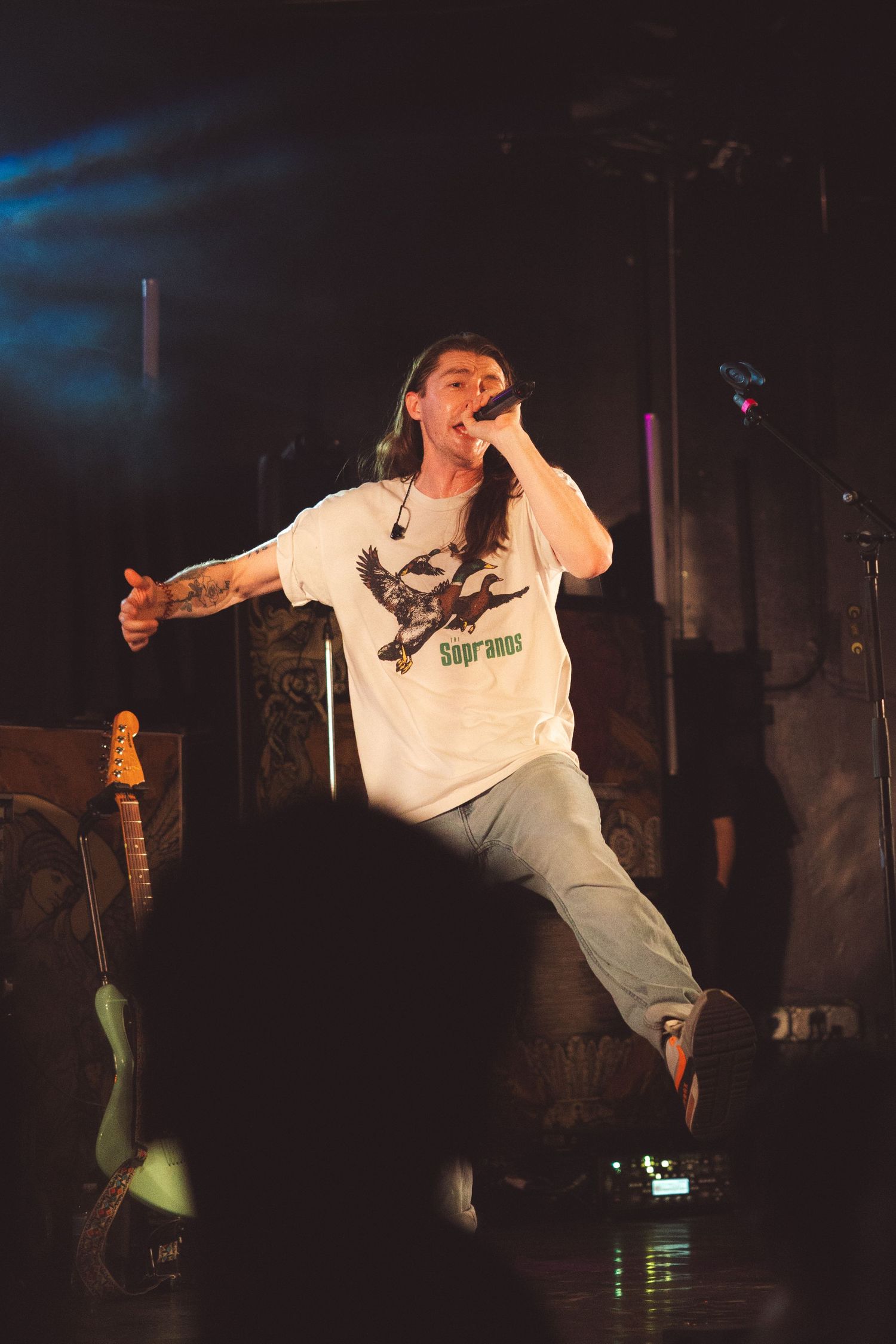 Performer in white t-shirt energetically dancing and jumping on dark stage during live performance.