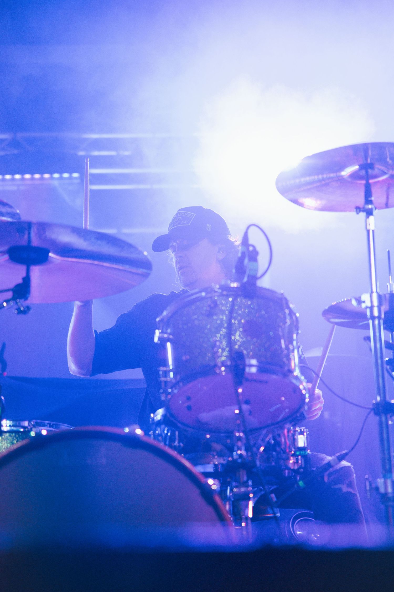 A drummer performs on stage surrounded by blue atmospheric lighting and fog during a live concert performance.