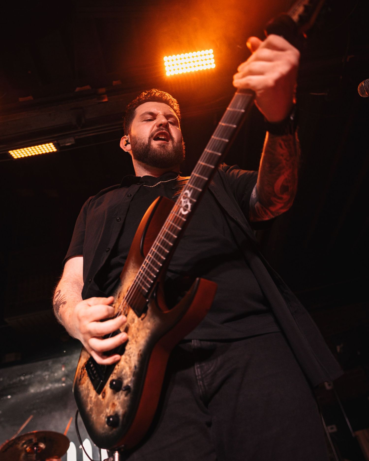 Tattooed male guitarist performing on stage with electric guitar under dramatic orange stage lighting.