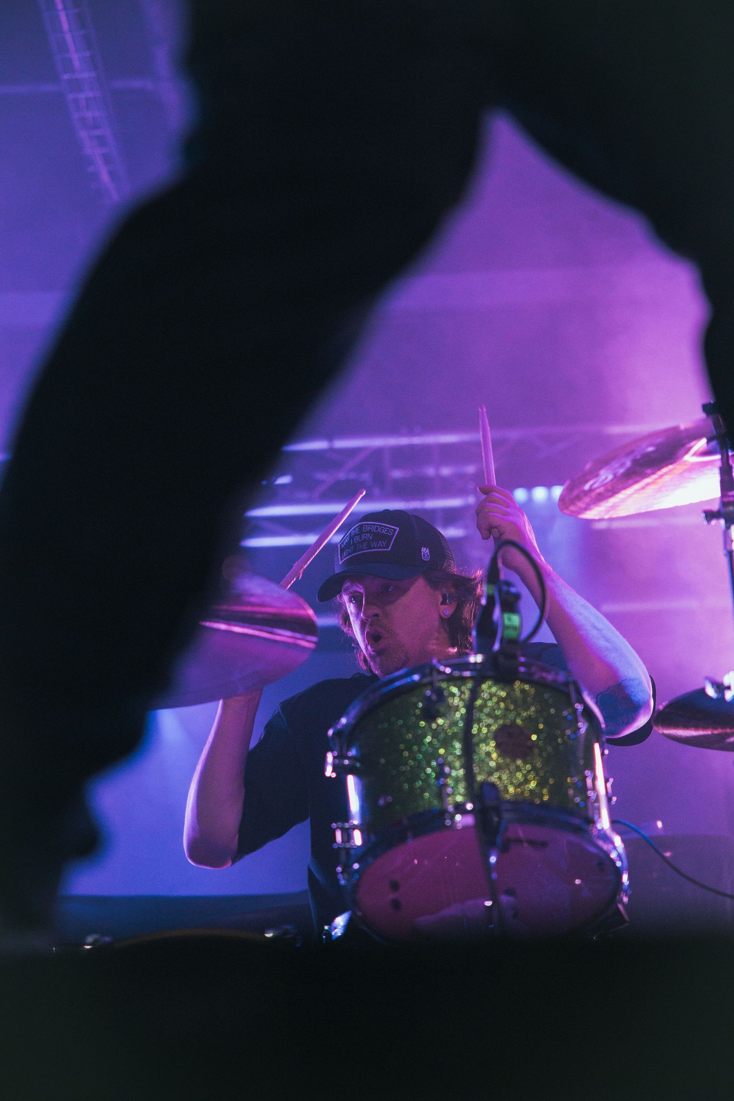 Drummer performing on stage illuminated by purple and blue stage lights in a dark concert venue.