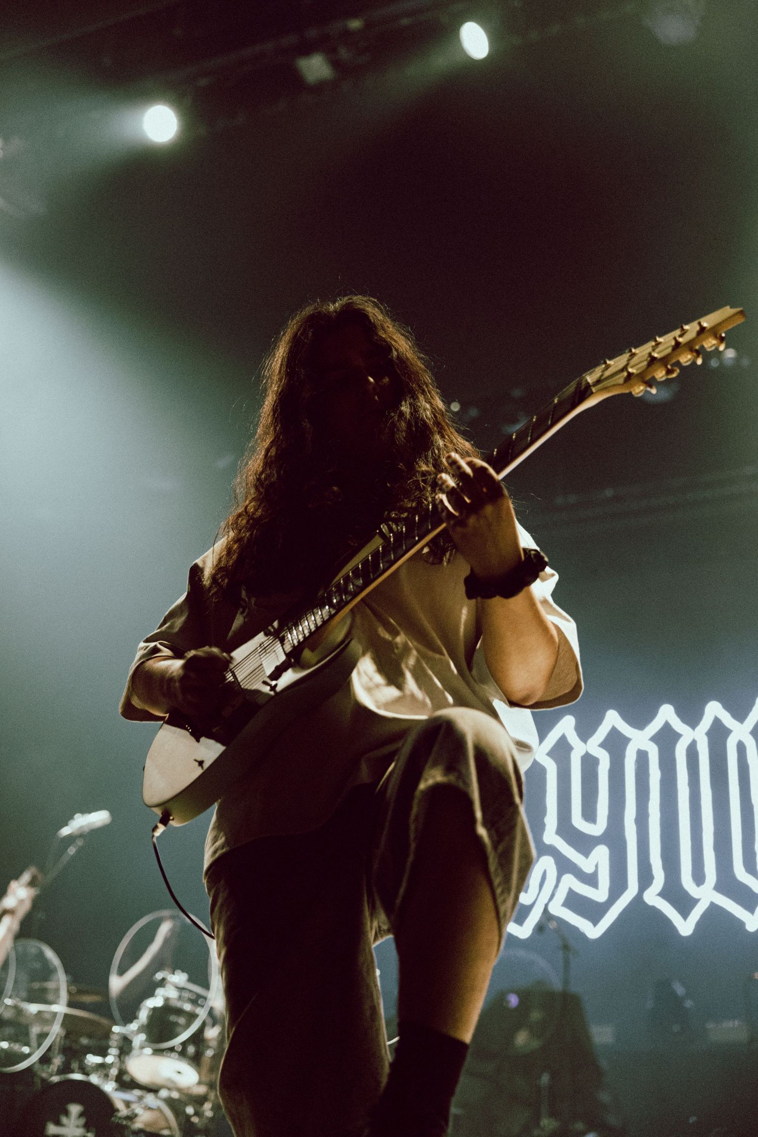Long-haired guitarist performing on stage under dramatic green lights at a live metal concert.