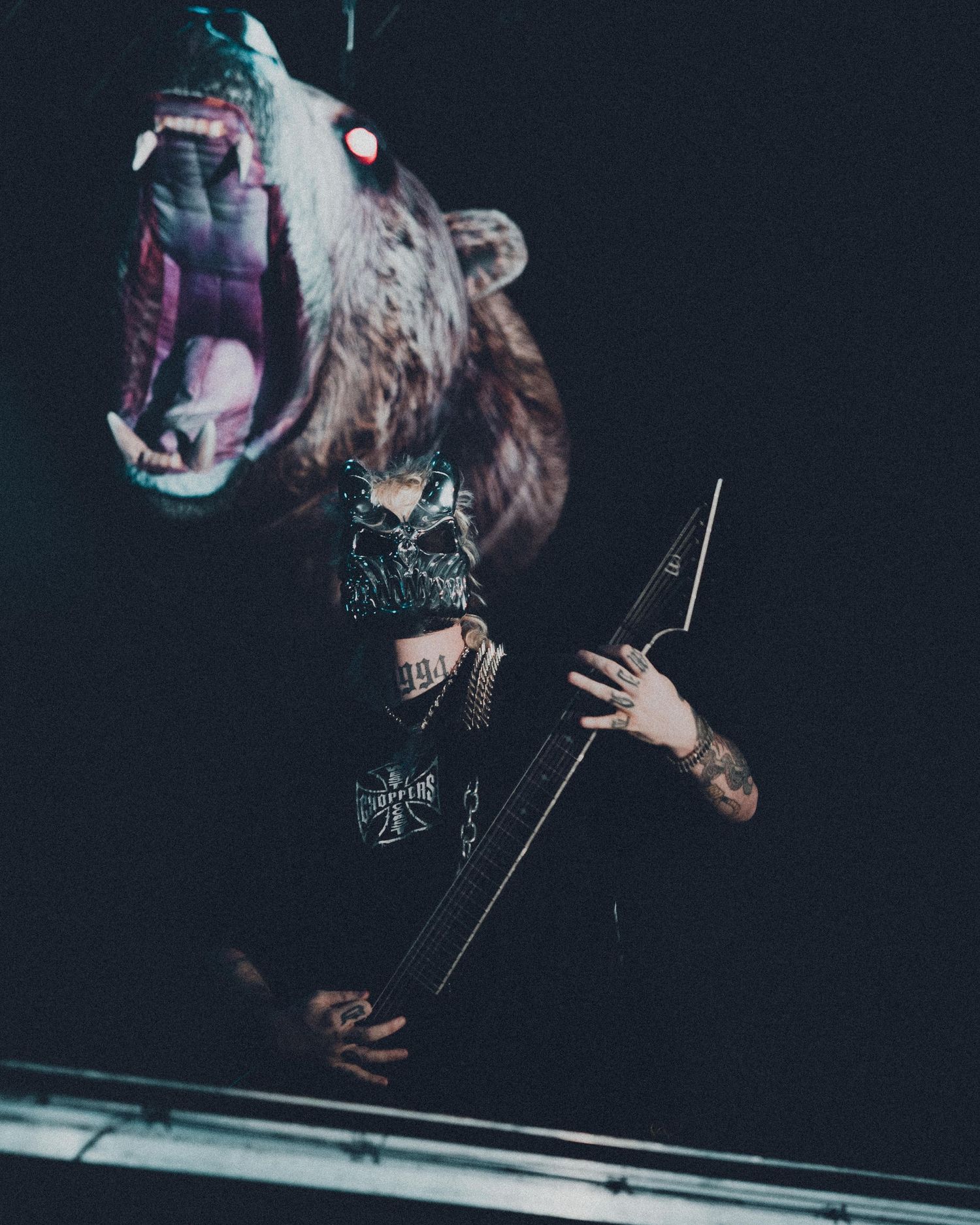 Tattooed musician plays dark guitar under a roaring bear mount in dramatic low-light setting.