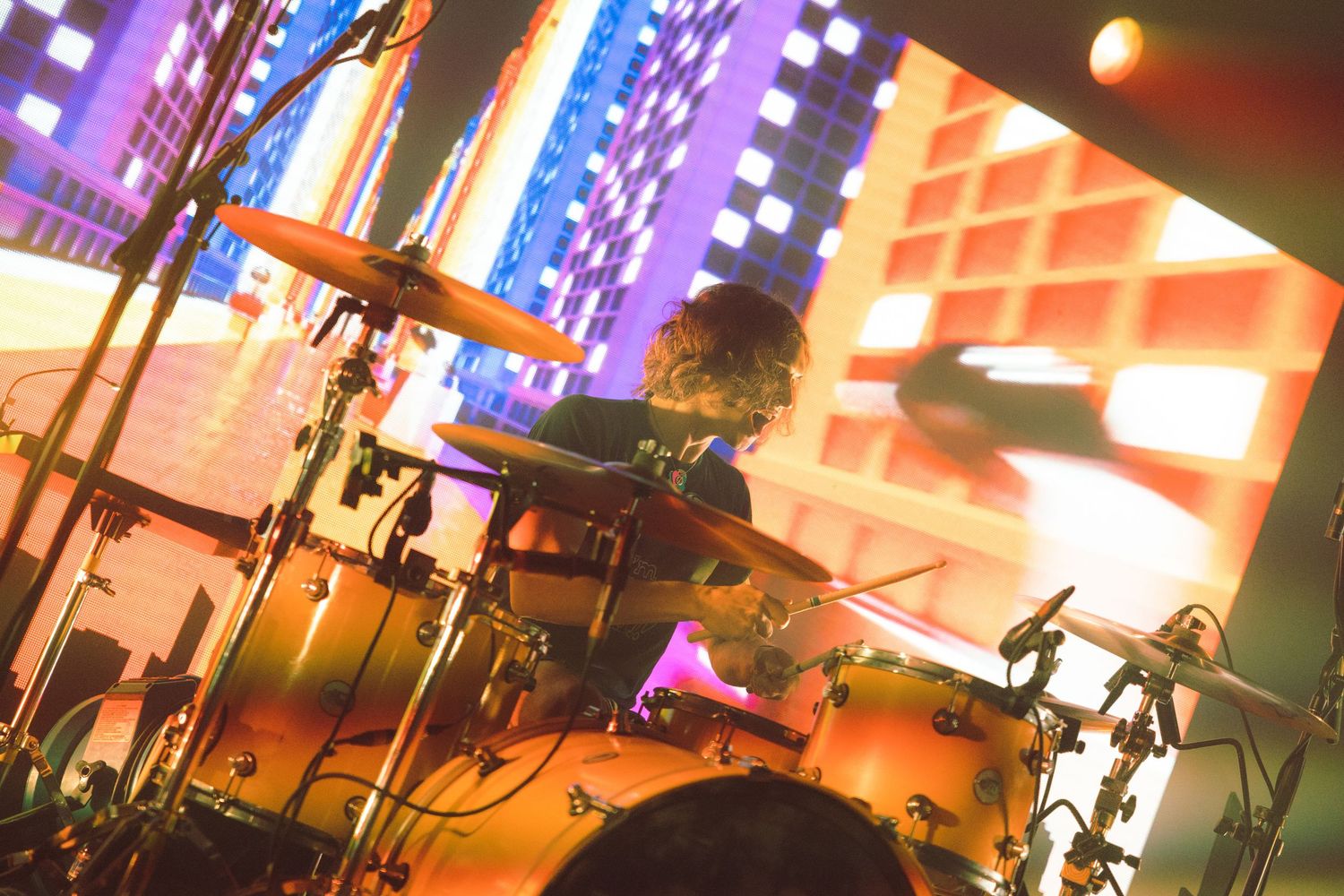 Drum kit setup on stage with colorful purple and orange lighting effects in the background during a live performance.