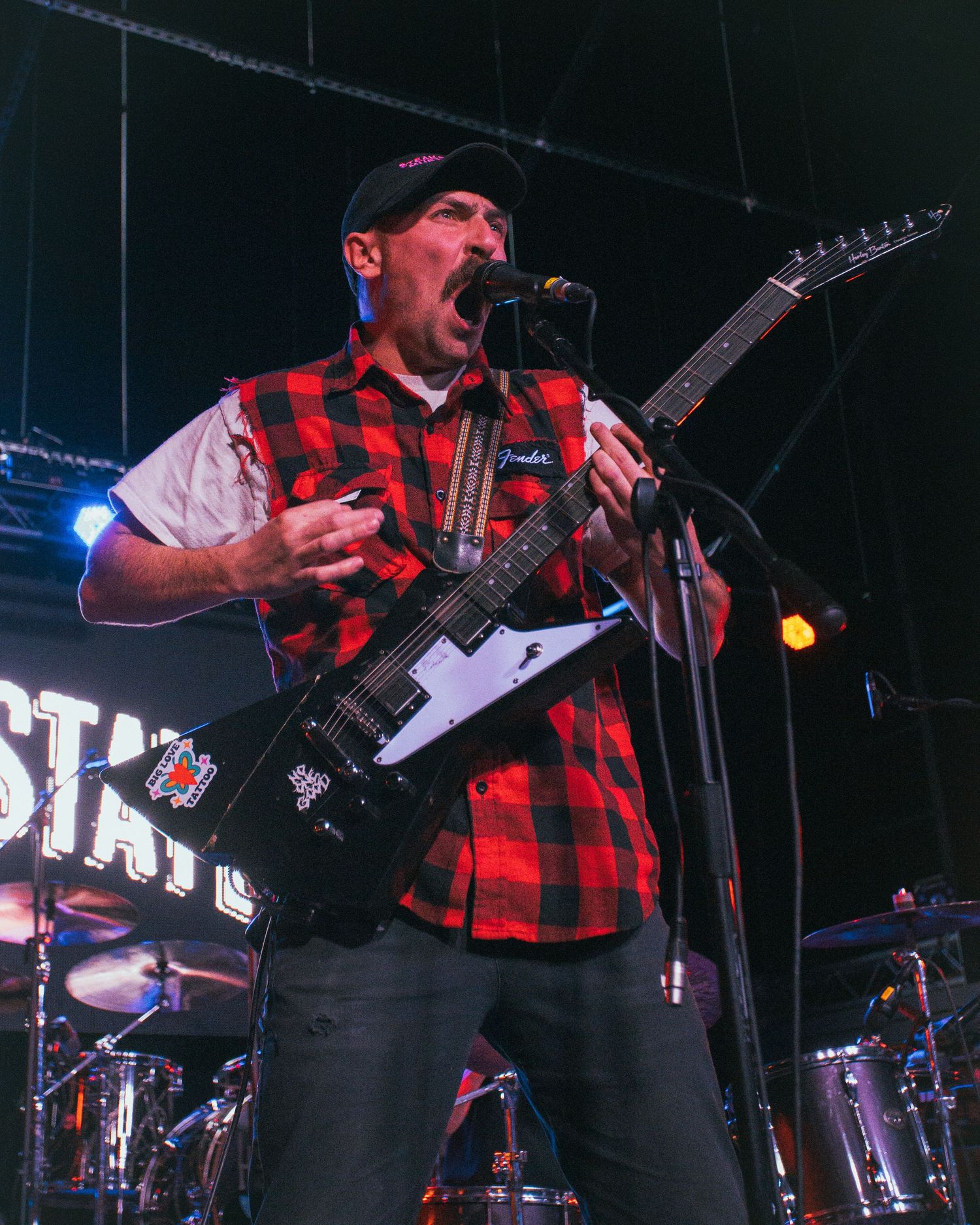 Musician in red plaid vest performs energetically with electric guitar on stage under colored lights.
