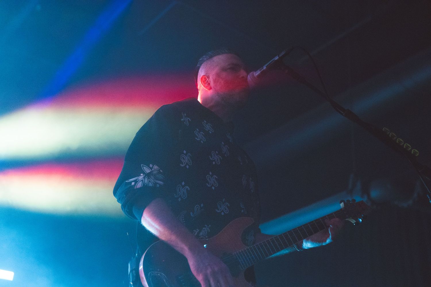 A musician plays guitar on stage with dramatic blue and pink concert lighting in the background.
