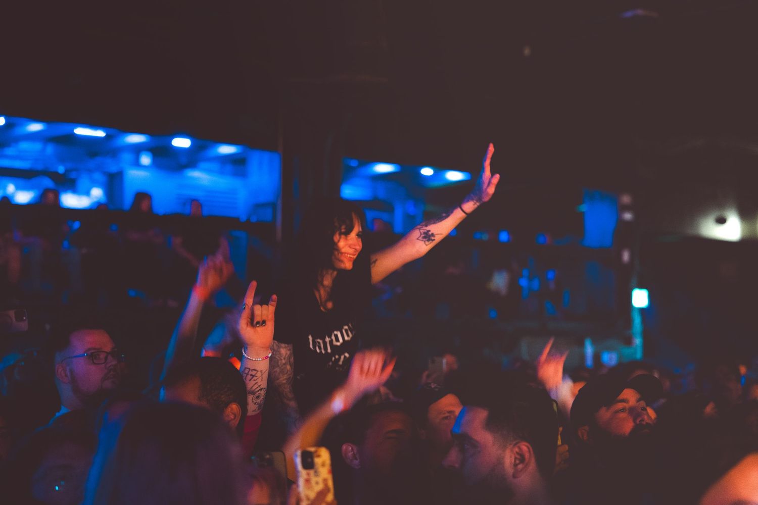 Concert crowd raises hands and rock horns in dark venue with blue lighting during live music performance.