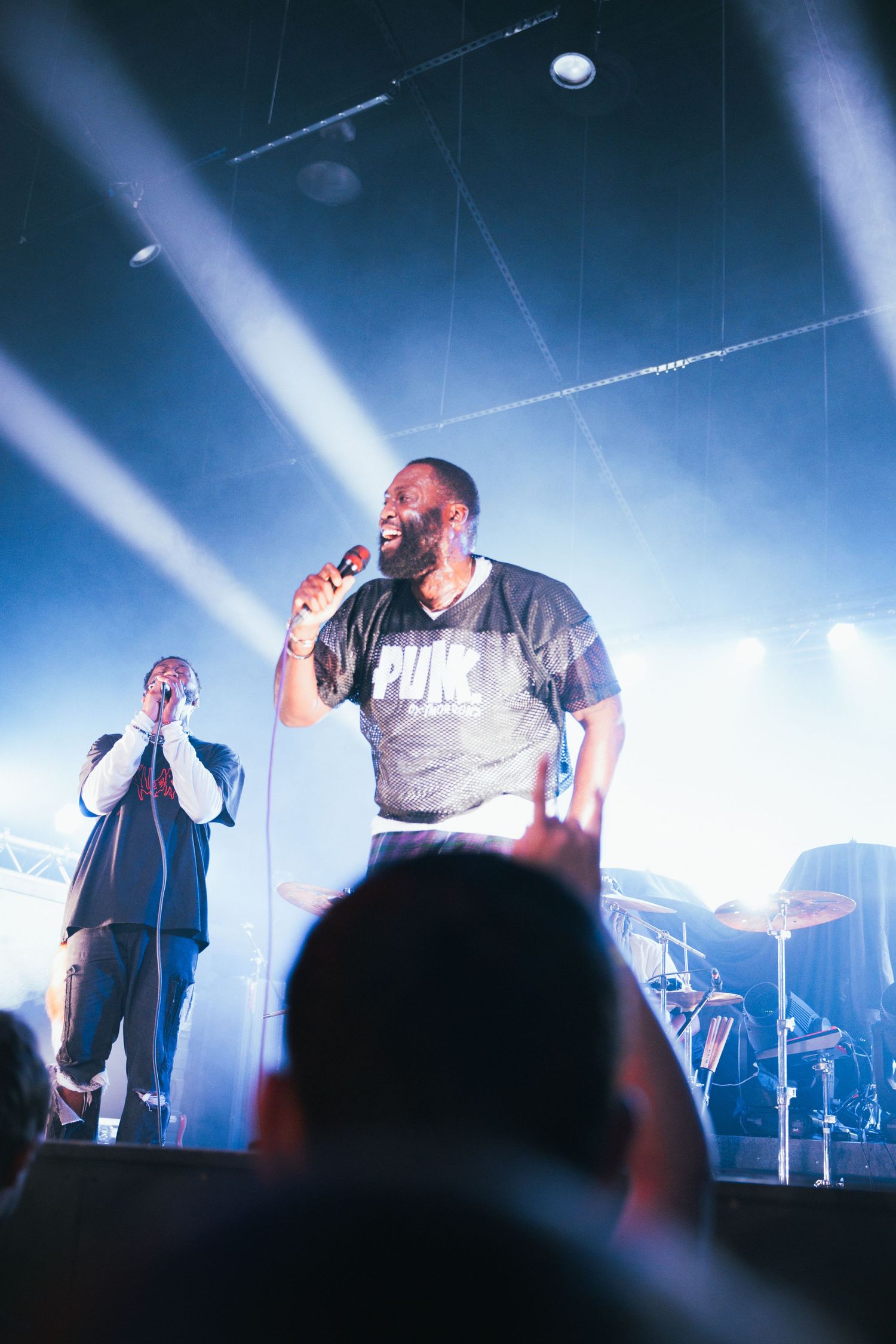 Performer on stage singing into microphone with dramatic blue lighting and silhouettes of band members in background.