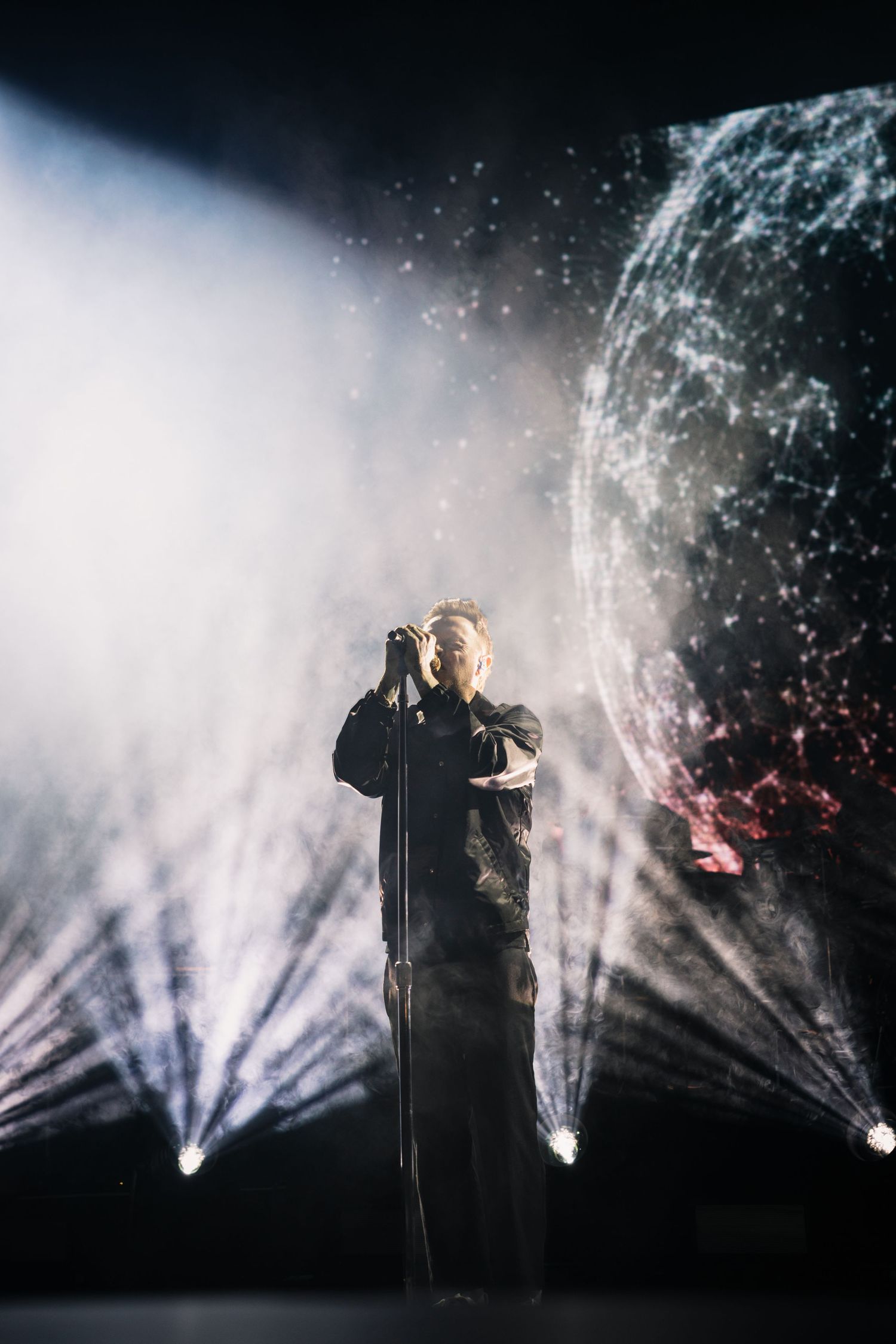 Singer performing on stage with dramatic light beams and glowing spherical visual display in dark concert venue.