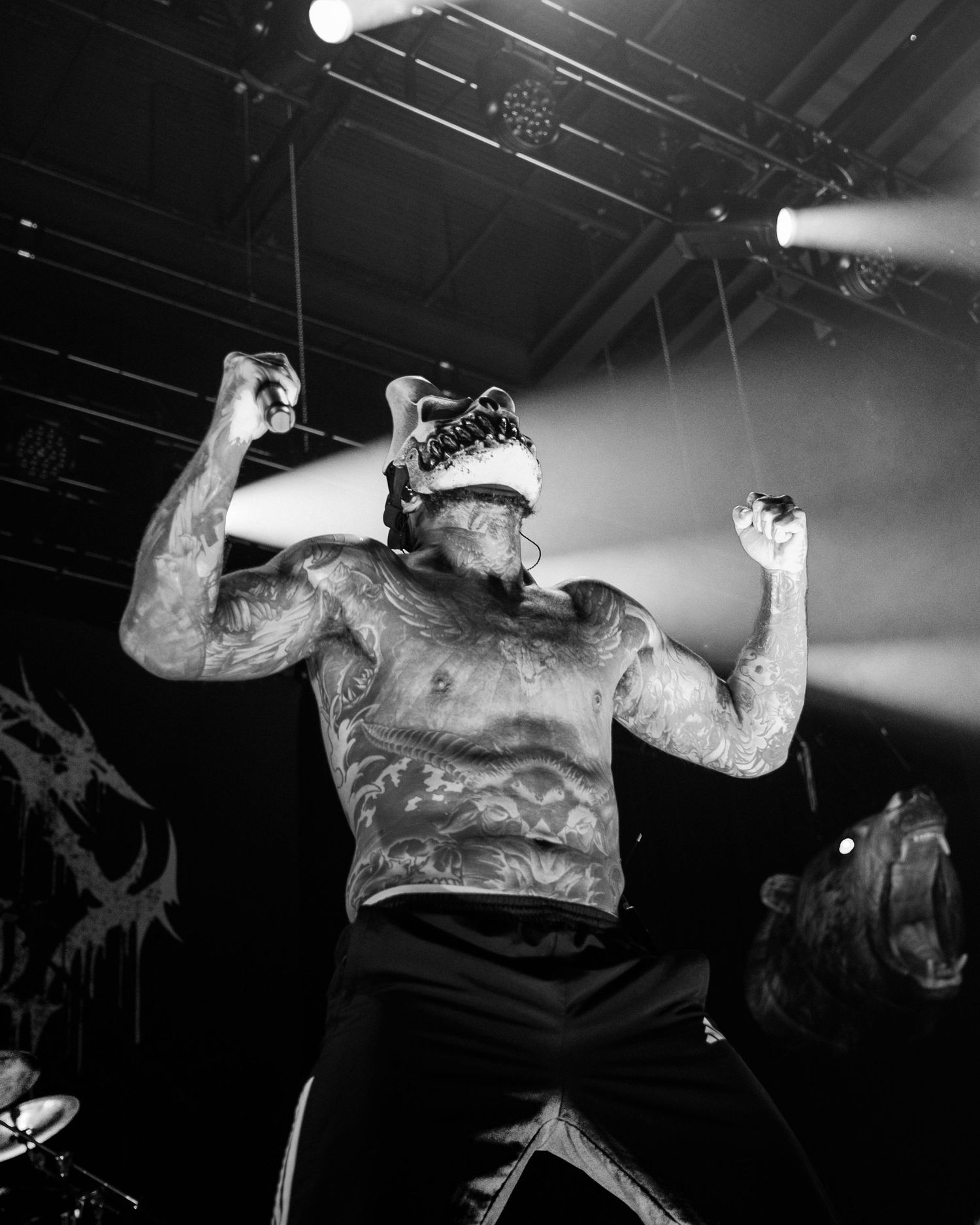 Shirtless tattooed metal performer on stage raising fists under dramatic lighting in black and white concert photo.