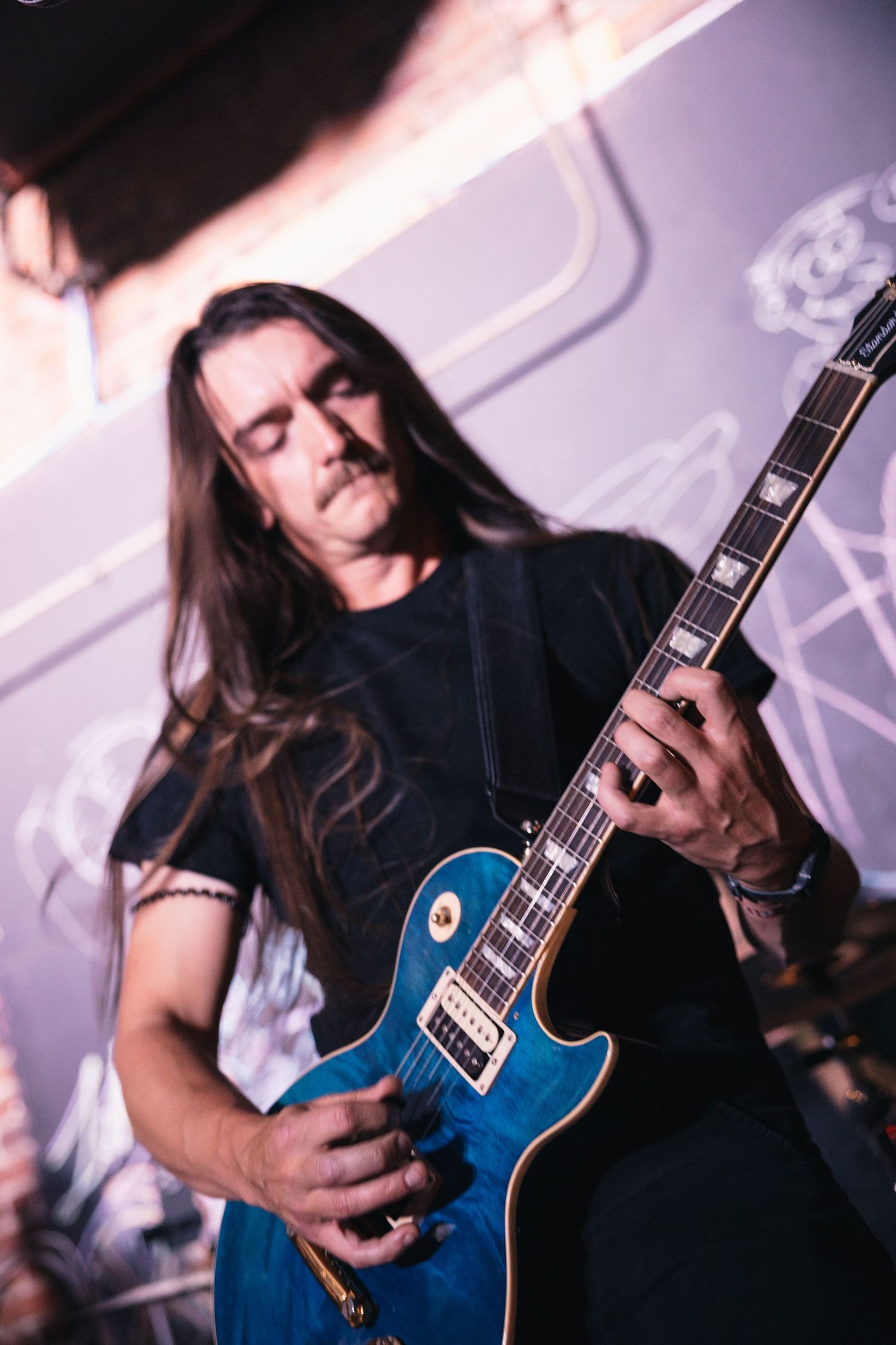 A musician plays a blue Les Paul guitar with long hair and eyes closed during a performance.