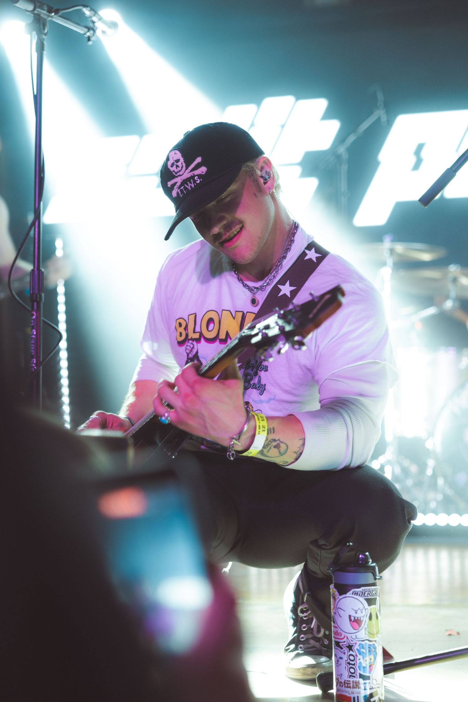 A guitarist in a black cap and Blonde shirt performs on stage under bright lights at a live concert.