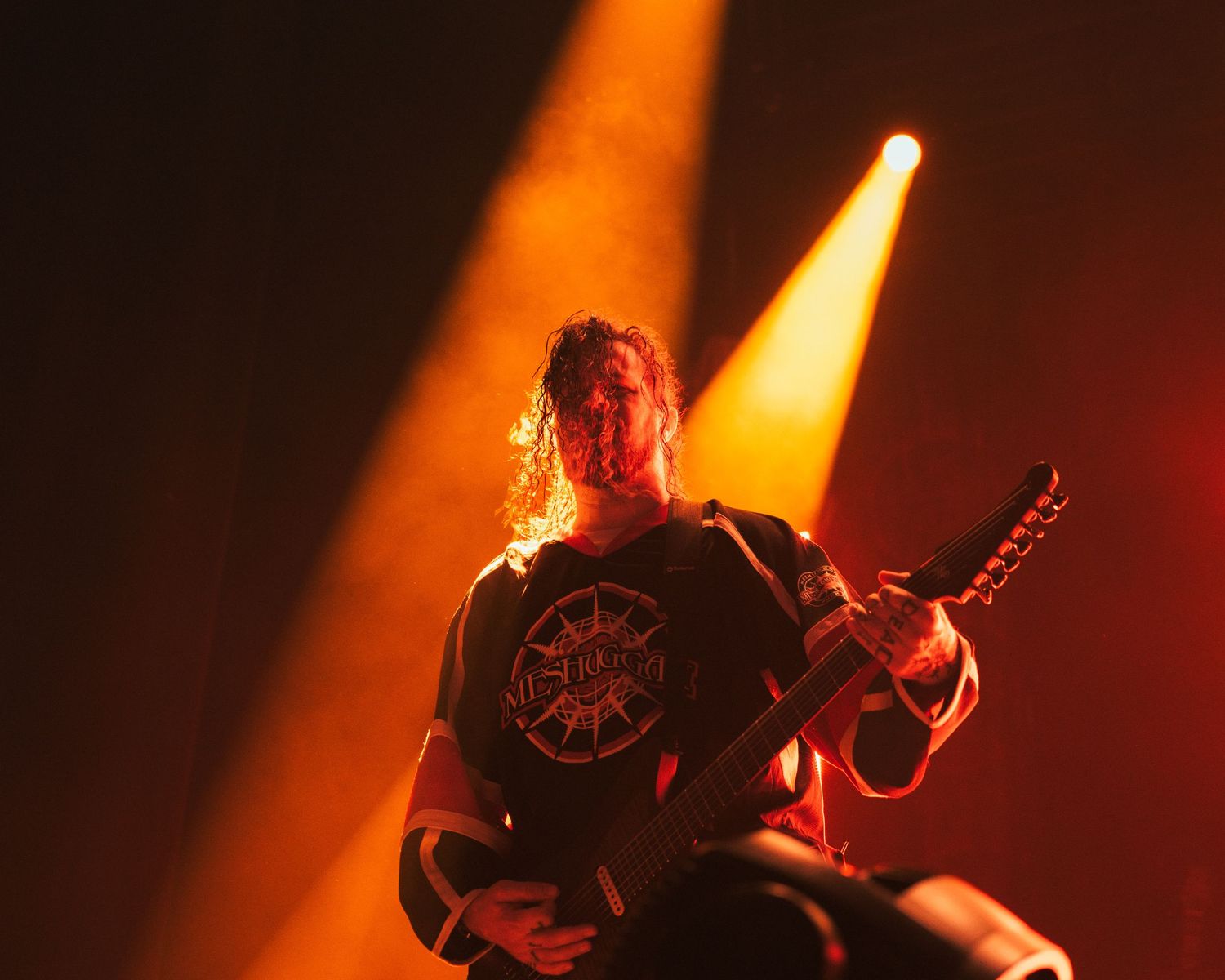 A guitarist performs on stage under dramatic orange and yellow spotlights, wearing a dark band t-shirt.