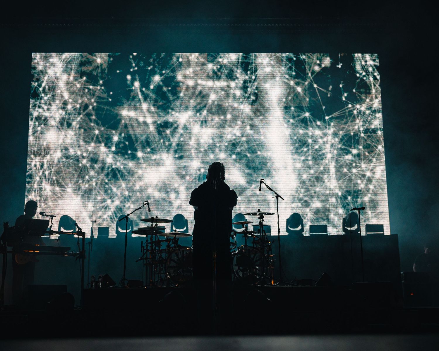 Silhouetted performer stands before a massive glowing neural network display on stage with drums and equipment visible.