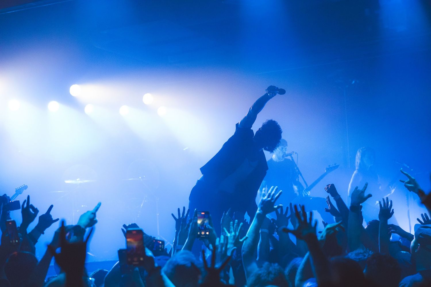 Silhouetted crowd enjoying a concert with raised hands against blue foggy lighting and stage performers.