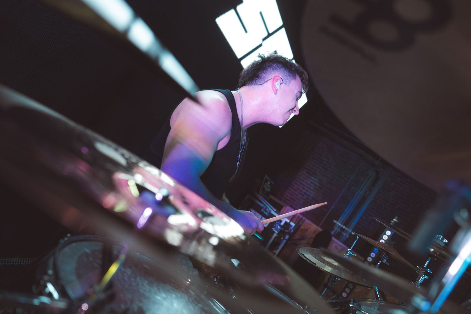 A drummer performs intensely on stage under colorful lights in a dark venue.