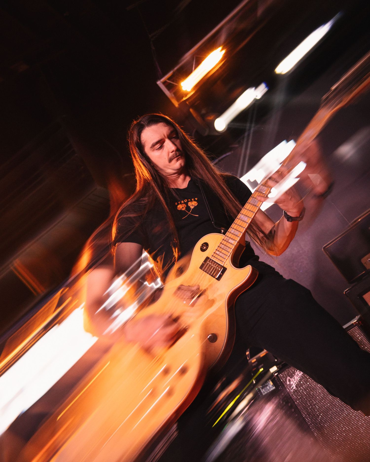 A guitarist performs on stage with motion blur effects and dramatic orange lighting creating an energetic atmosphere.