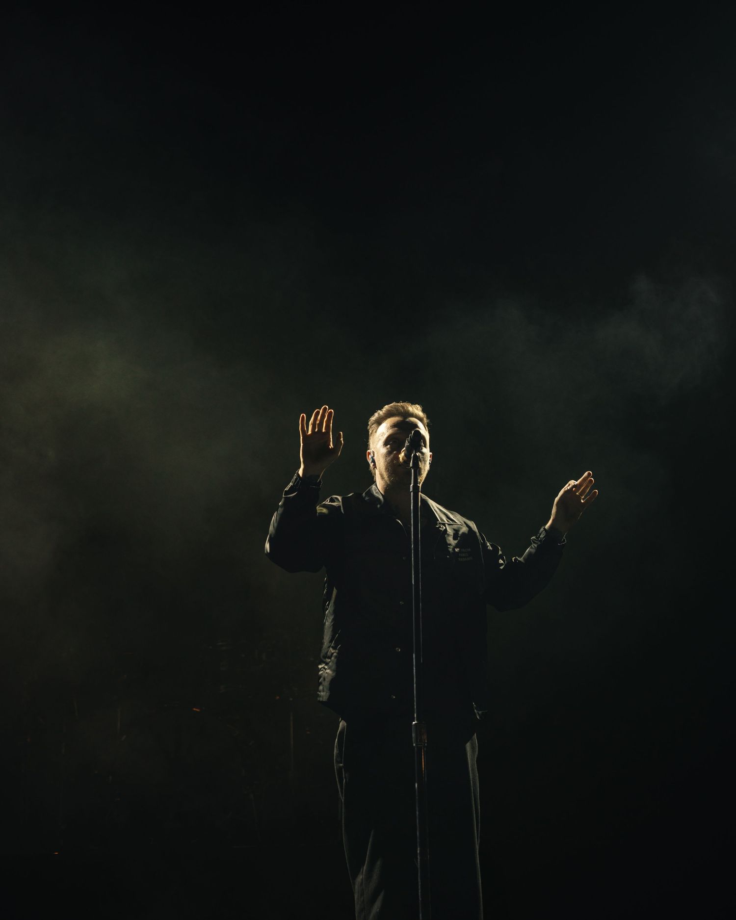 A dramatic performer stands on a dark stage with hands raised, illuminated by moody backlighting and atmospheric smoke.