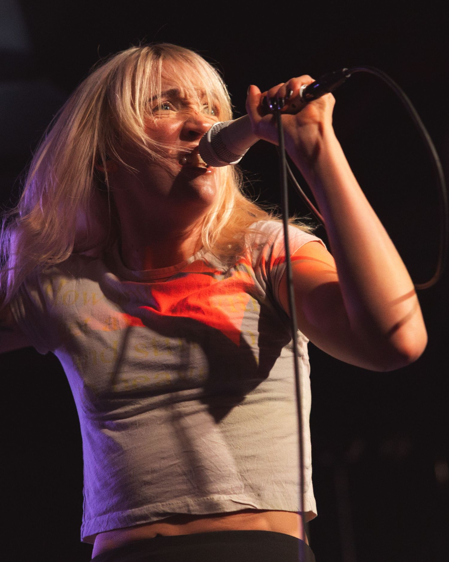 A performer sings passionately on stage in a white shirt against a dark background with dramatic purple lighting.