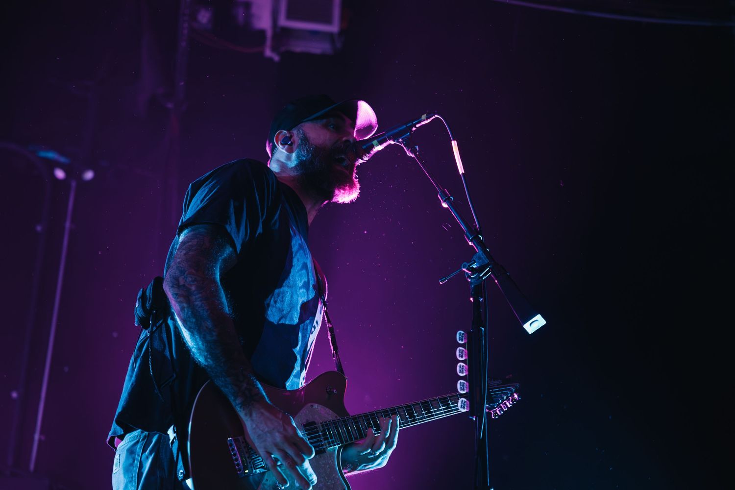 Guitarist performing on stage under purple lights, leaning into microphone while playing electric guitar at live concert.
