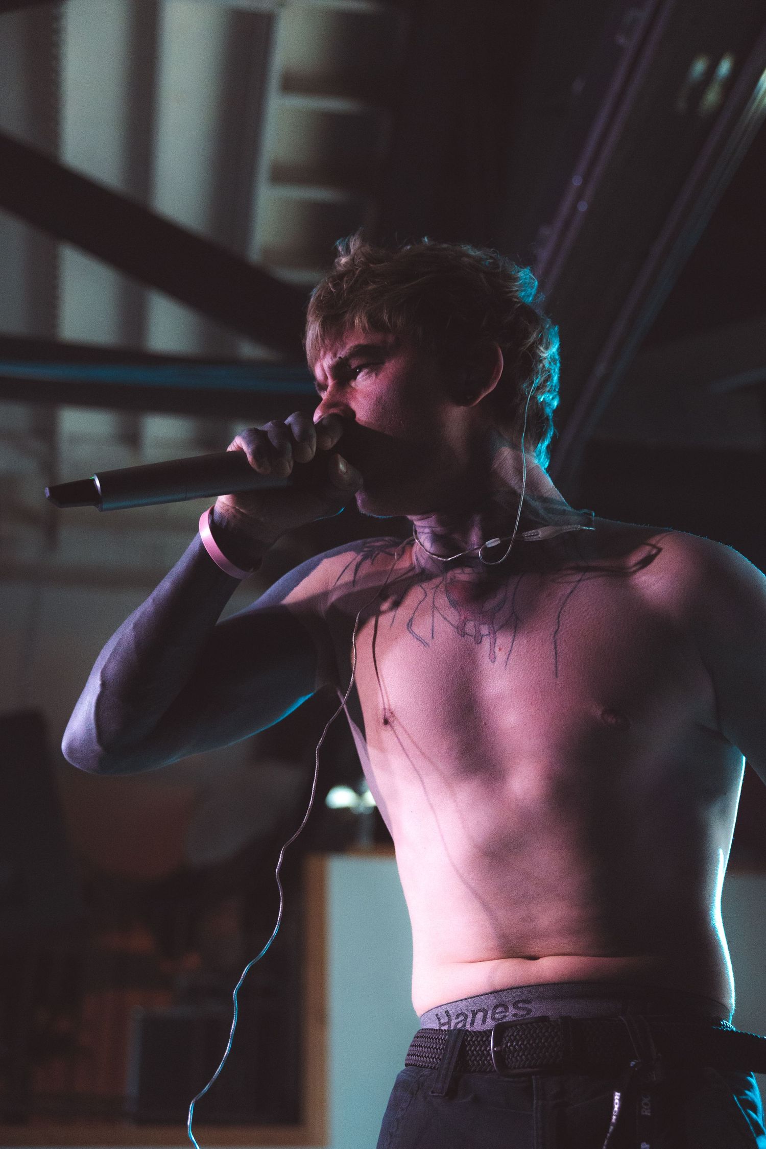 Shirtless performer sings passionately into microphone on dimly lit stage with dramatic blue lighting effects.