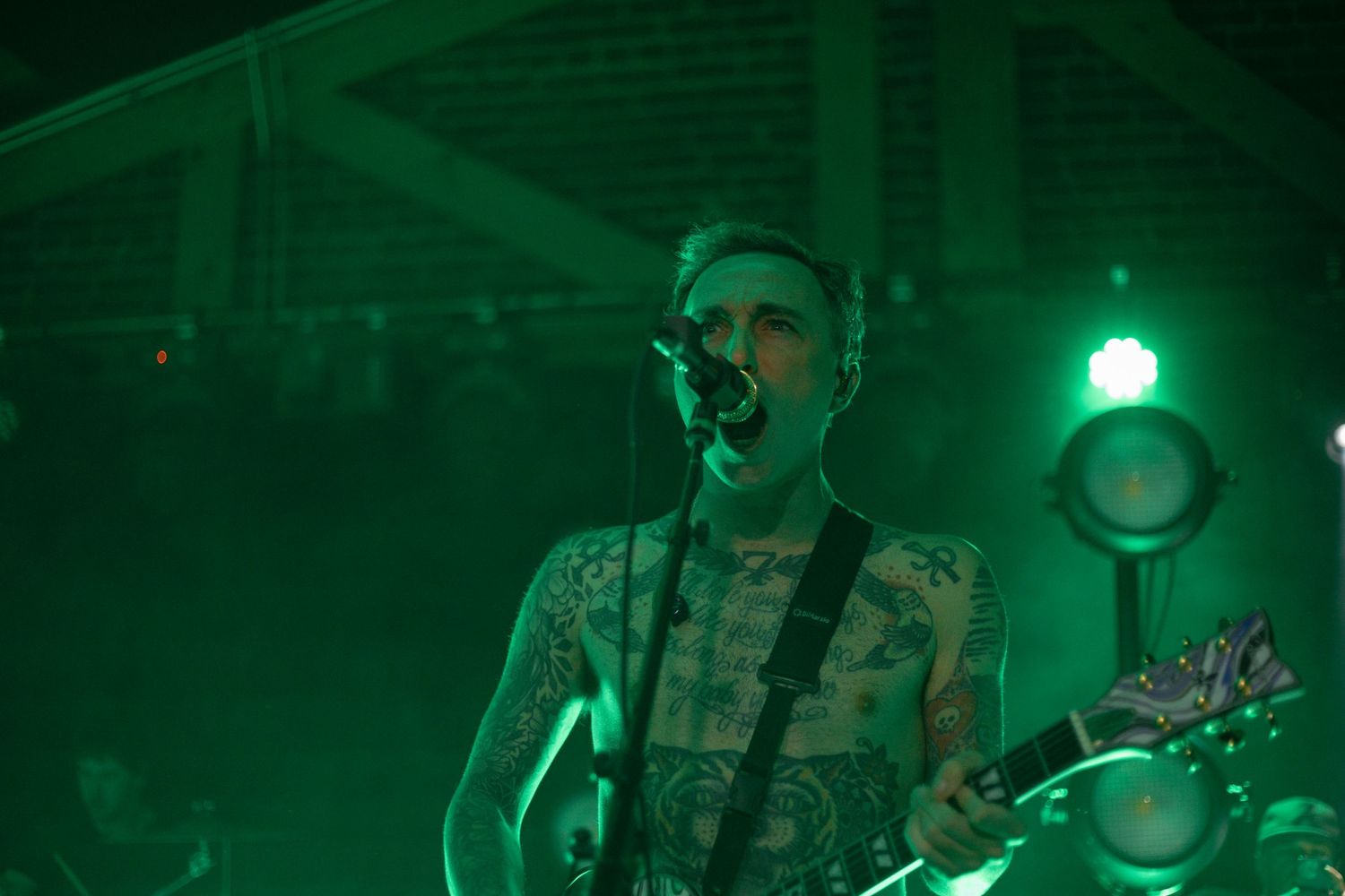 Musician performing with guitar under green stage lighting with visible tattoos.