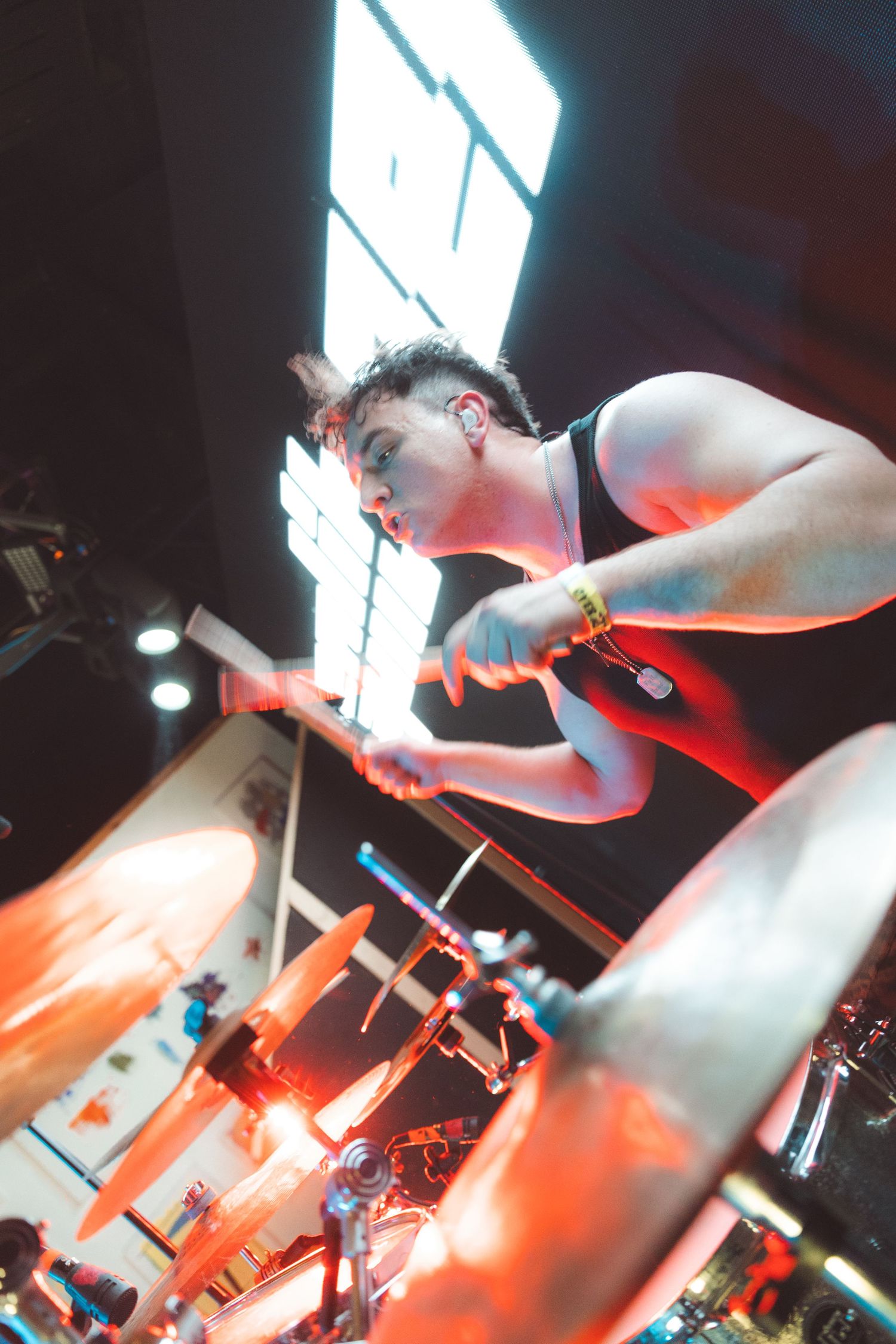 A drummer performing energetically on stage, captured from a low angle with dramatic red lighting and motion blur.