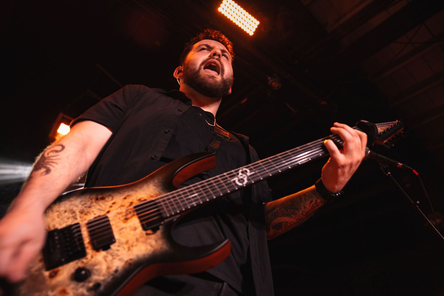 Tattooed male guitarist playing a multi-string bass guitar on stage under bright lights during a live concert.
