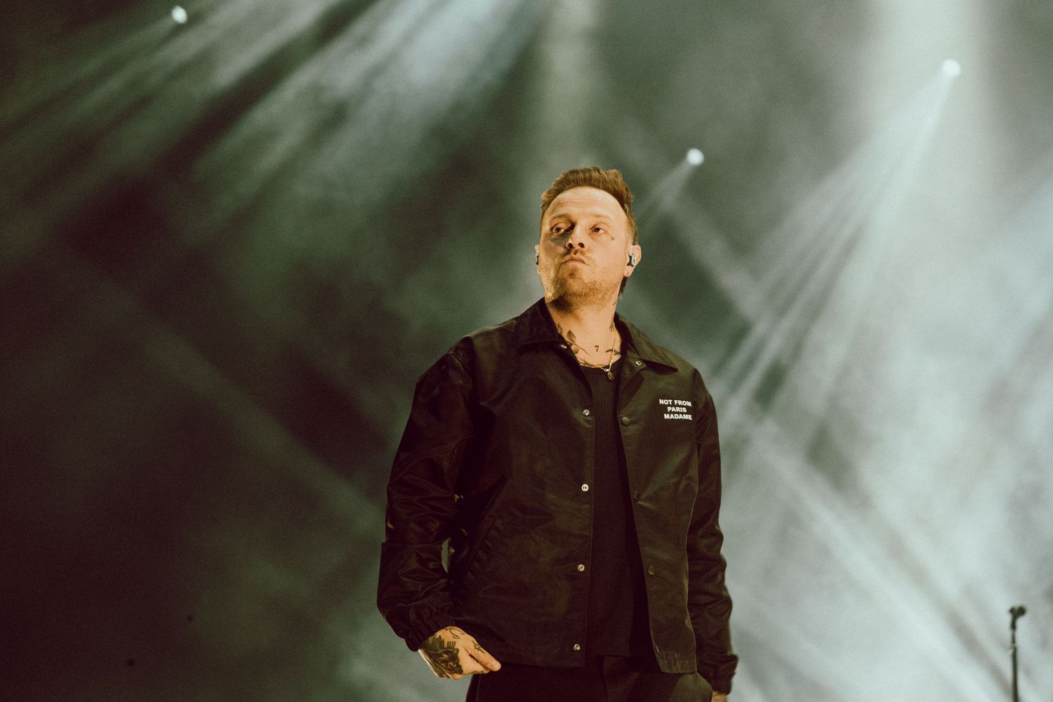 A male performer in a black jacket stands on stage under dramatic white spotlights.