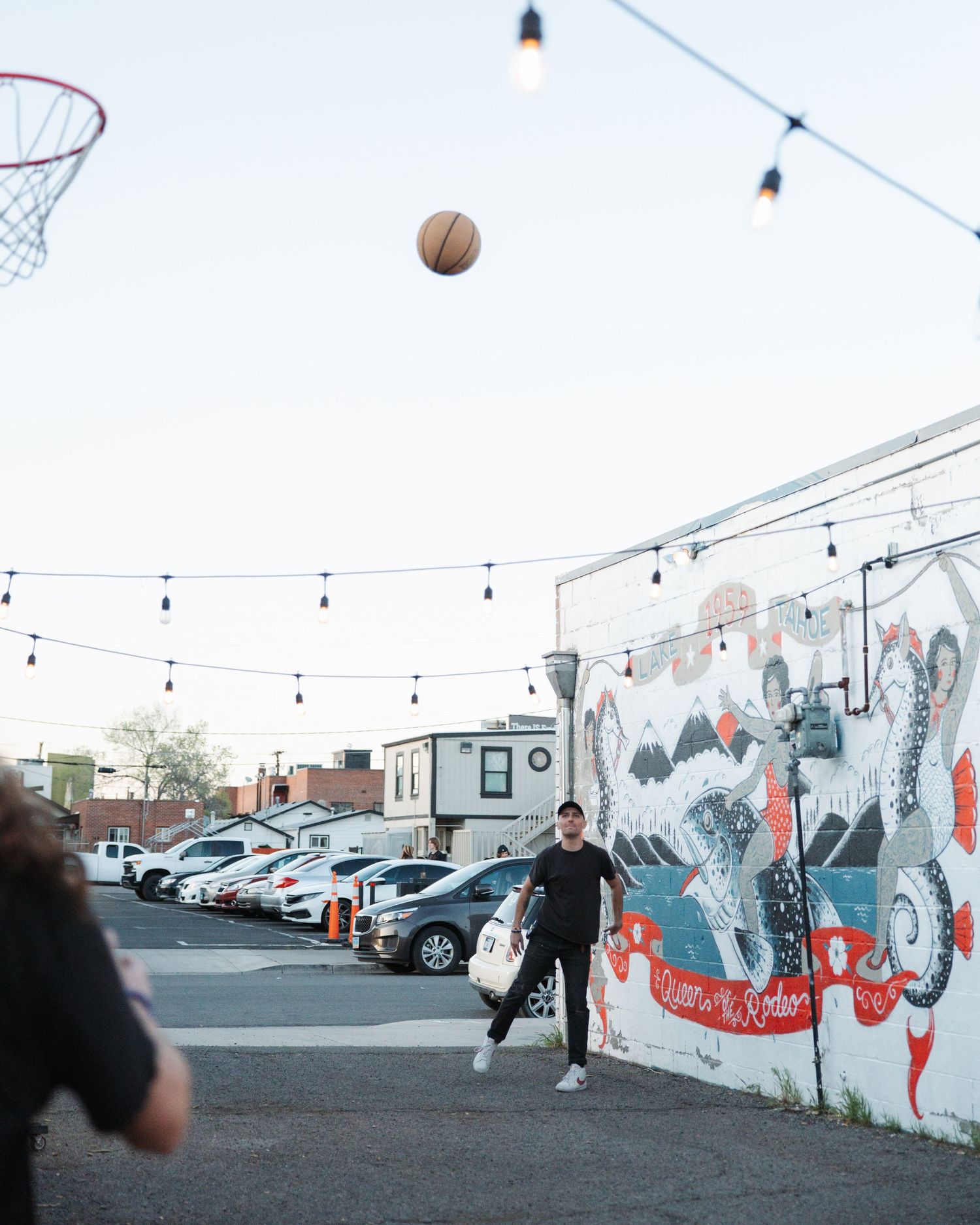 Person playing basketball under string lights in parking lot with mural wall backdrop.