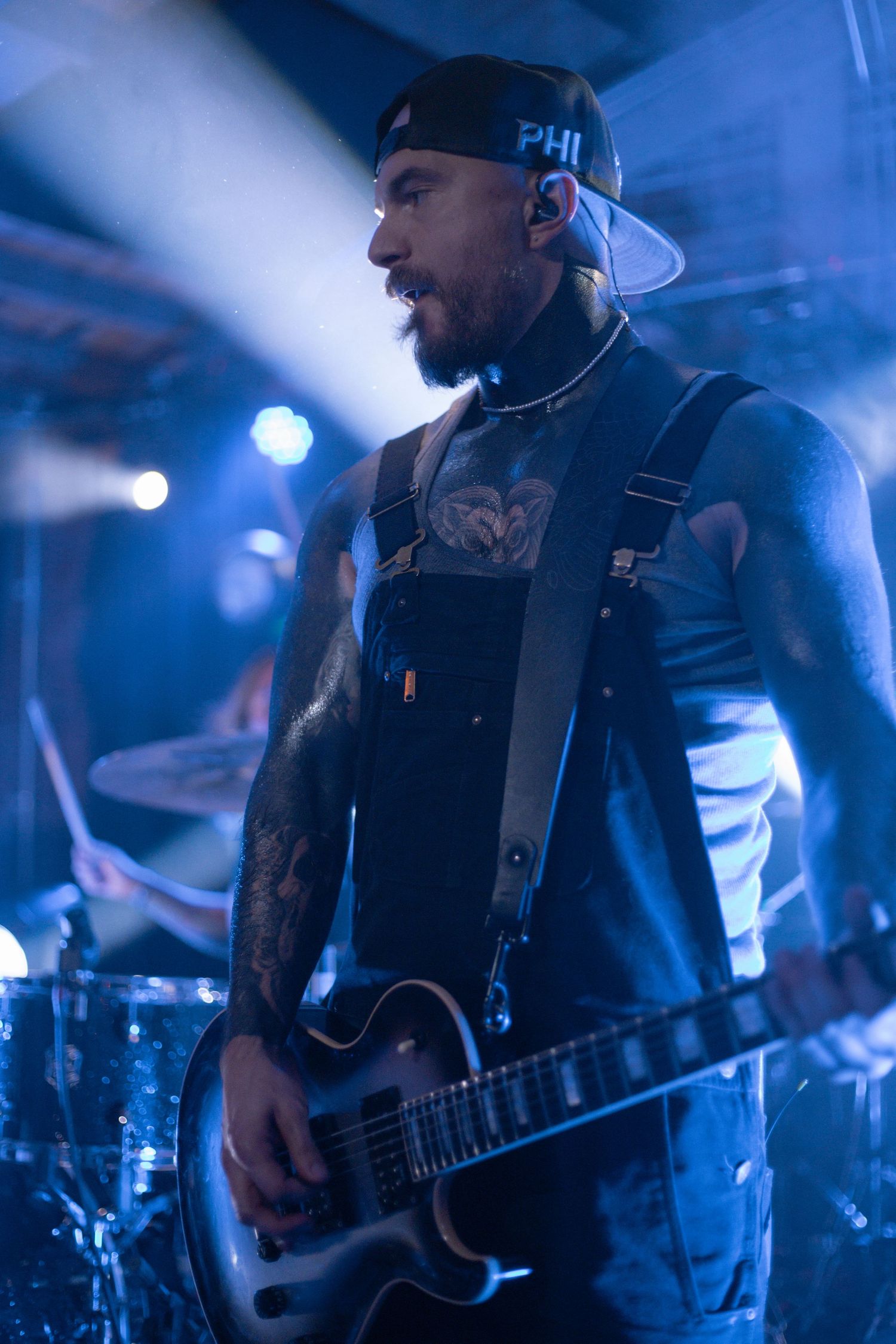 Tattooed musician plays guitar under dramatic blue stage lighting wearing backwards cap.