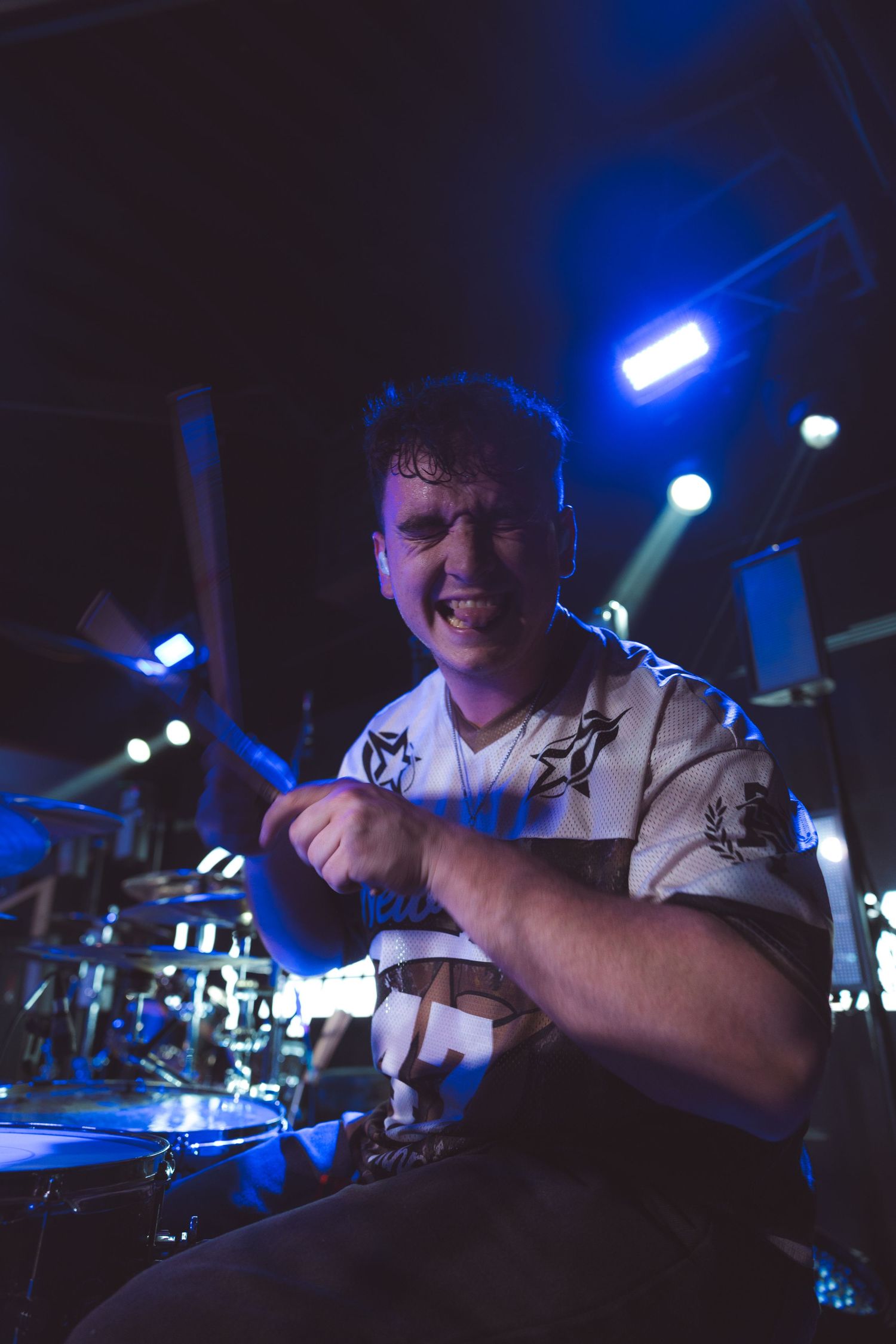 Energetic drummer performing on stage under blue lights, smiling while playing drums at a live concert.