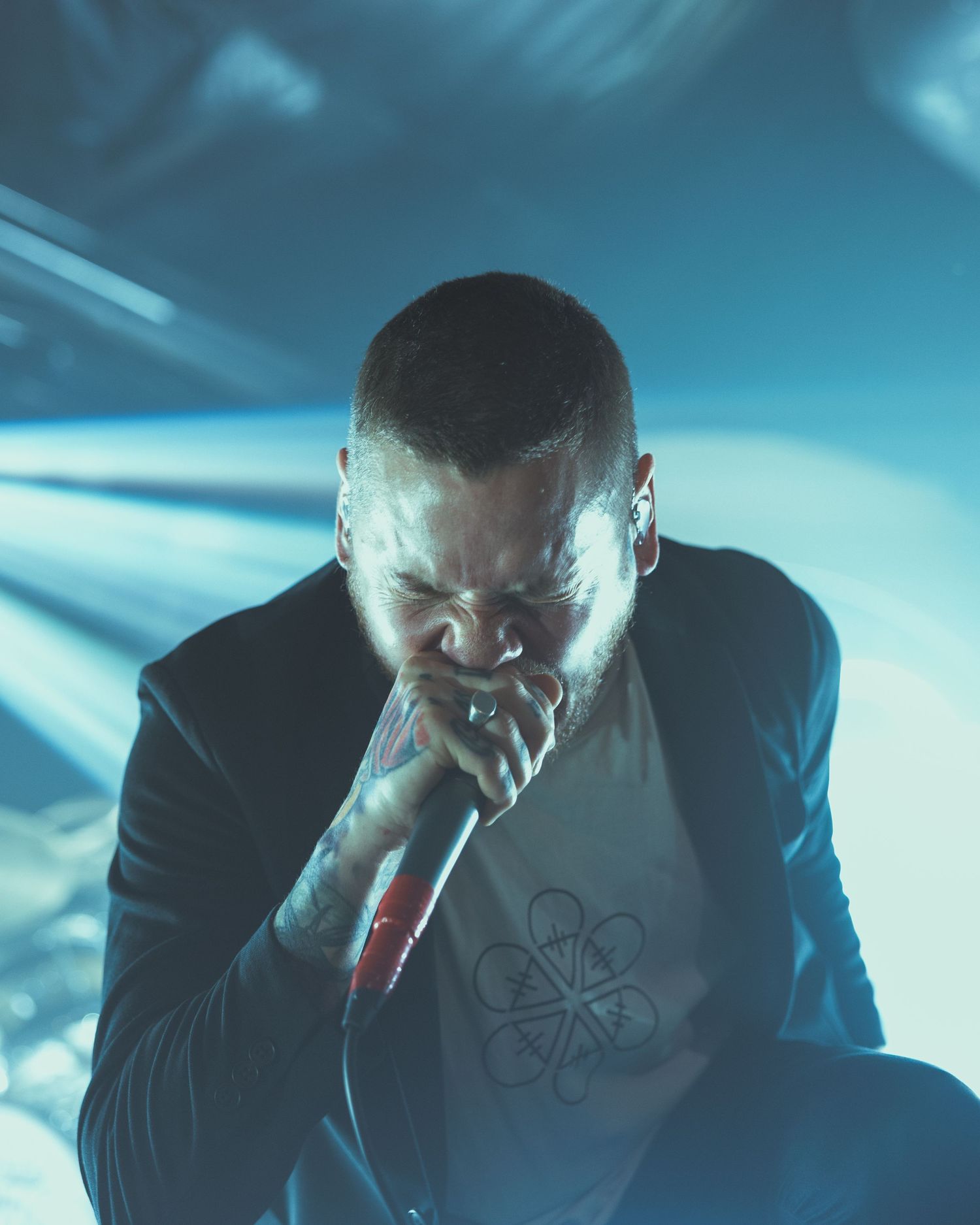 A bald male performer crouches on stage holding a microphone, wearing a black blazer and graphic tee under blue stage lighting.