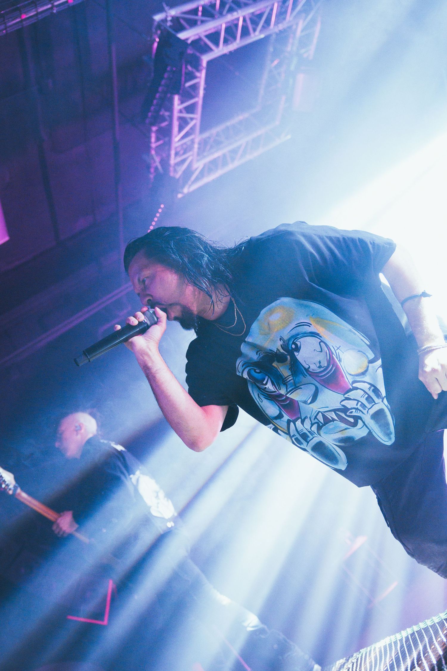 Performer in a black t-shirt singing on stage with dramatic blue lighting and metal truss rigging visible above.