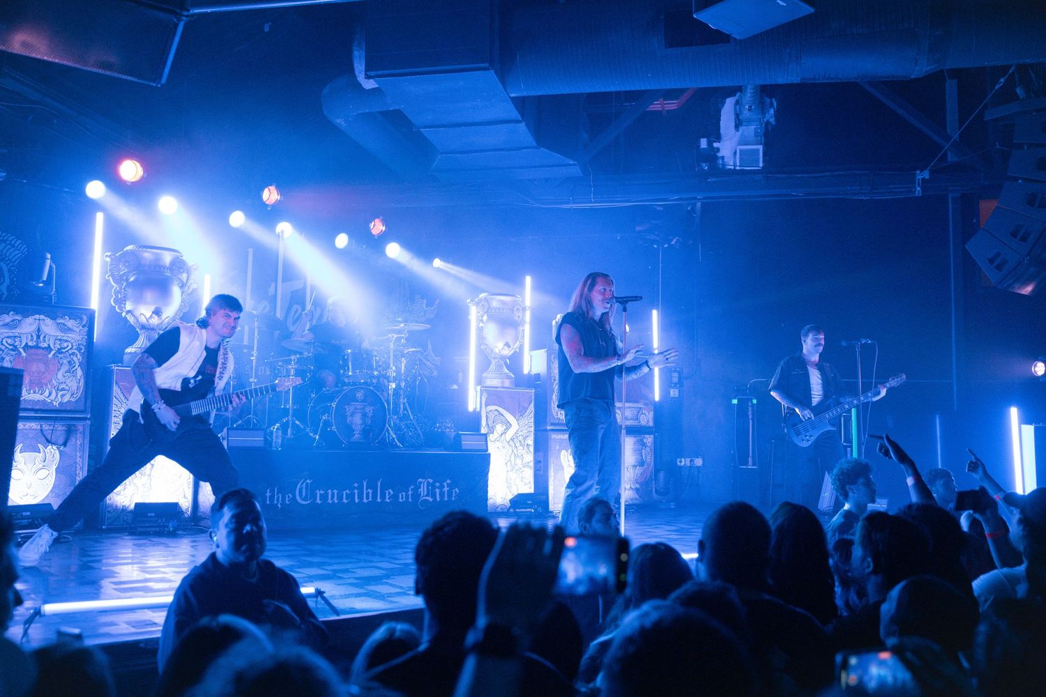 A concert stage bathed in dramatic blue lighting shows musicians performing for an energetic crowd at a live music venue.