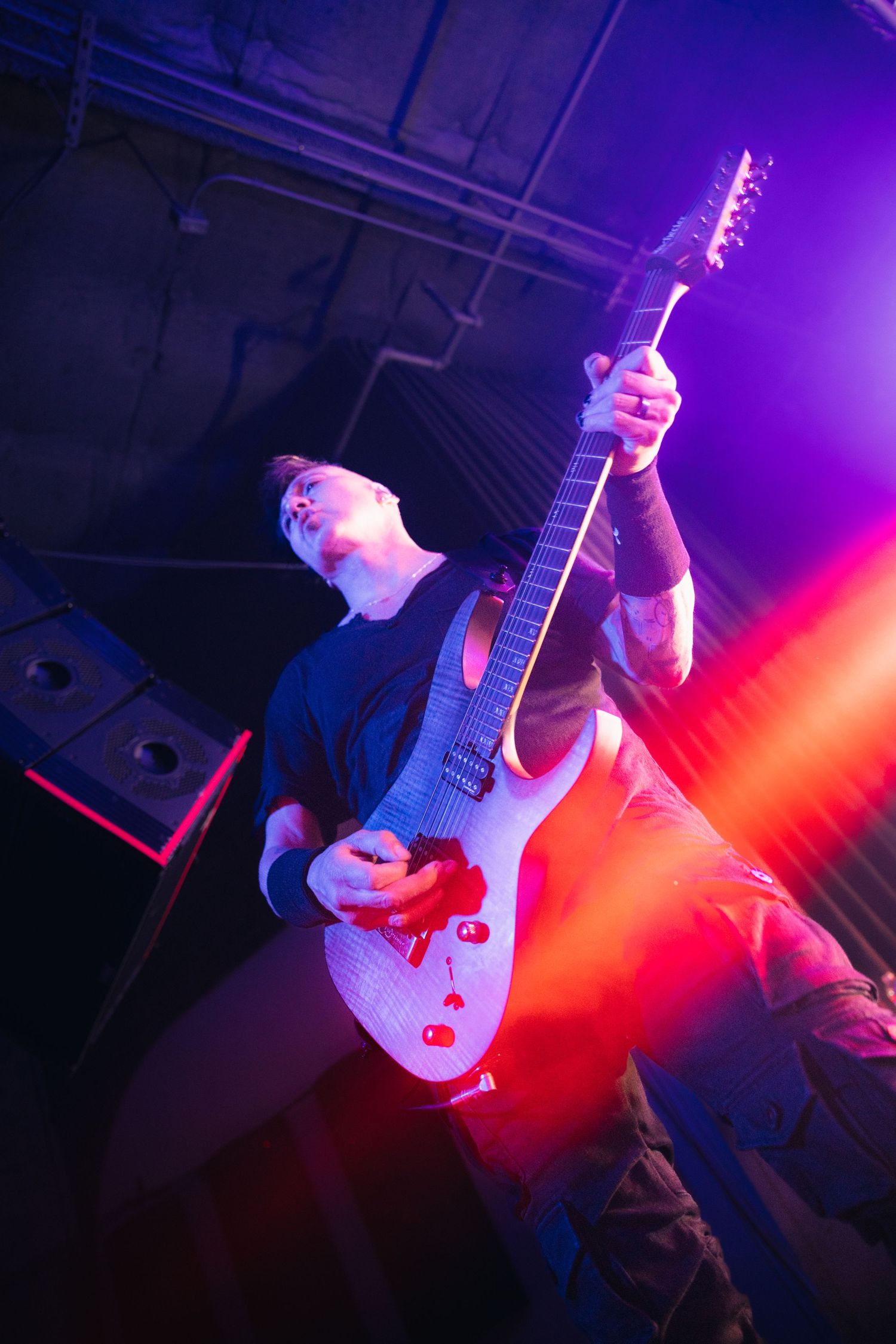 Guitarist performs on stage with white guitar under dramatic purple and red stage lighting at a live concert.