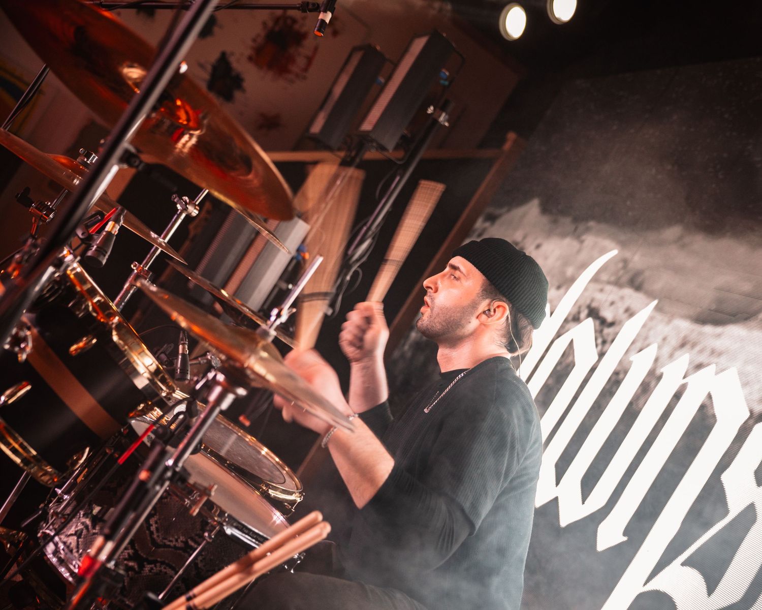 A drummer performs energetically on stage at a live metal concert, surrounded by cymbals and drums in dramatic lighting.