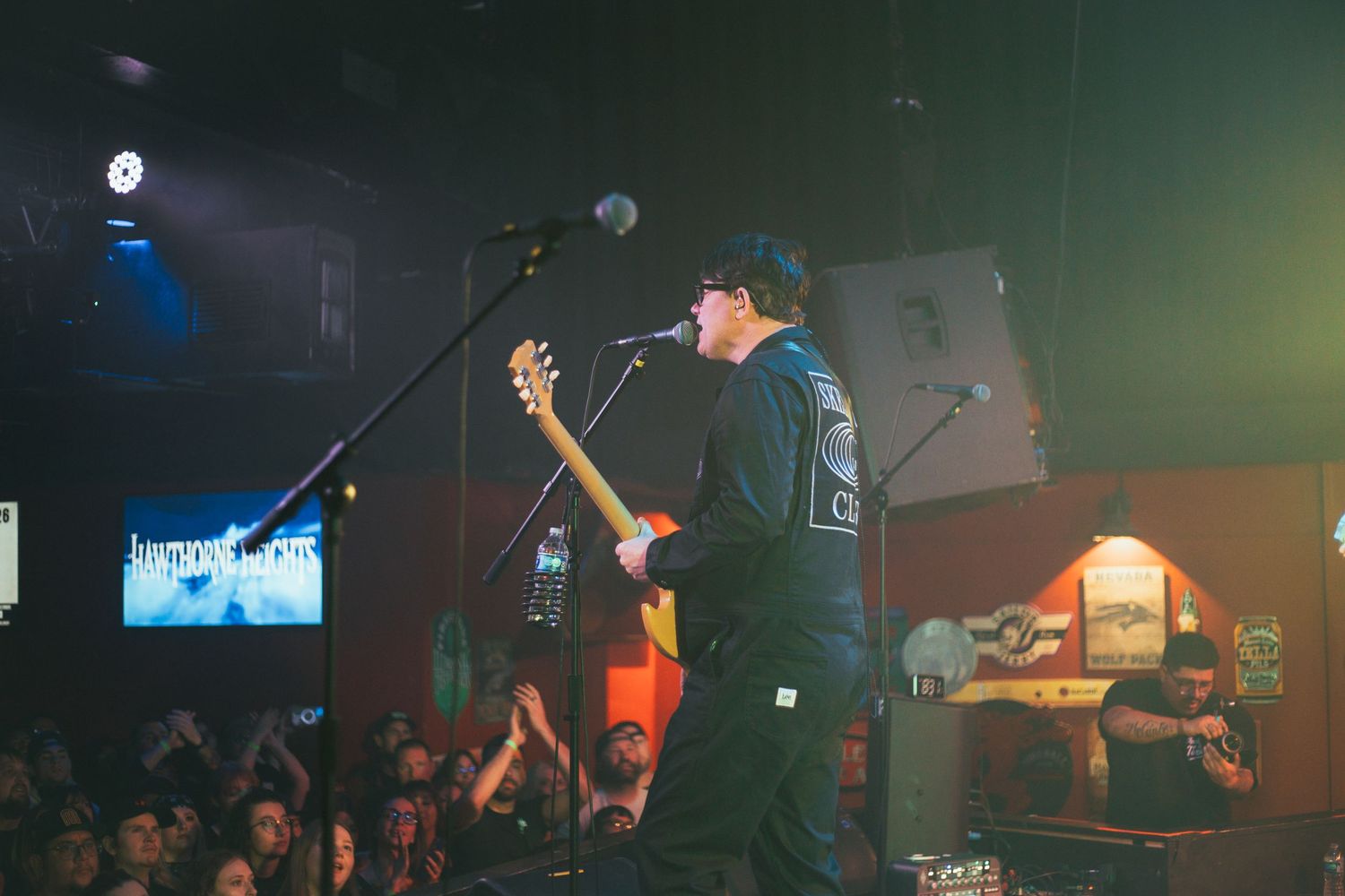 Musician performing with bass guitar on dimly lit stage with crowd visible.