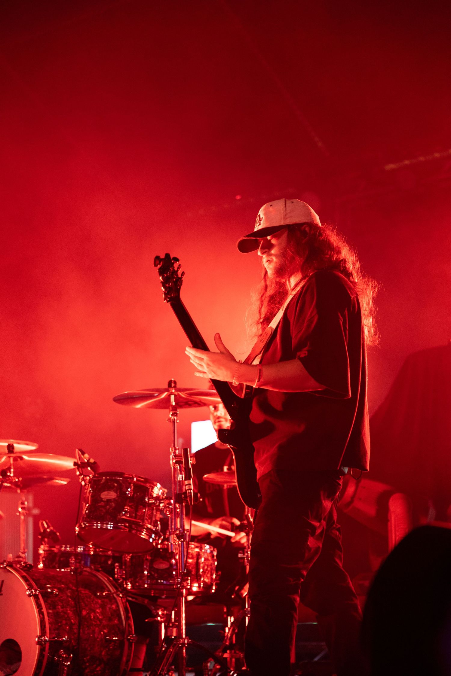 A musician performs on stage with a bass guitar bathed in dramatic red lighting next to a drum kit during a concert.