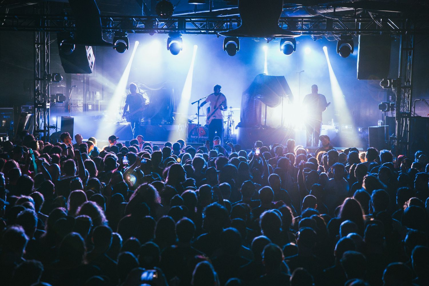 Live concert with a large crowd and band performing on a bright, blue-lit stage in an indoor venue.