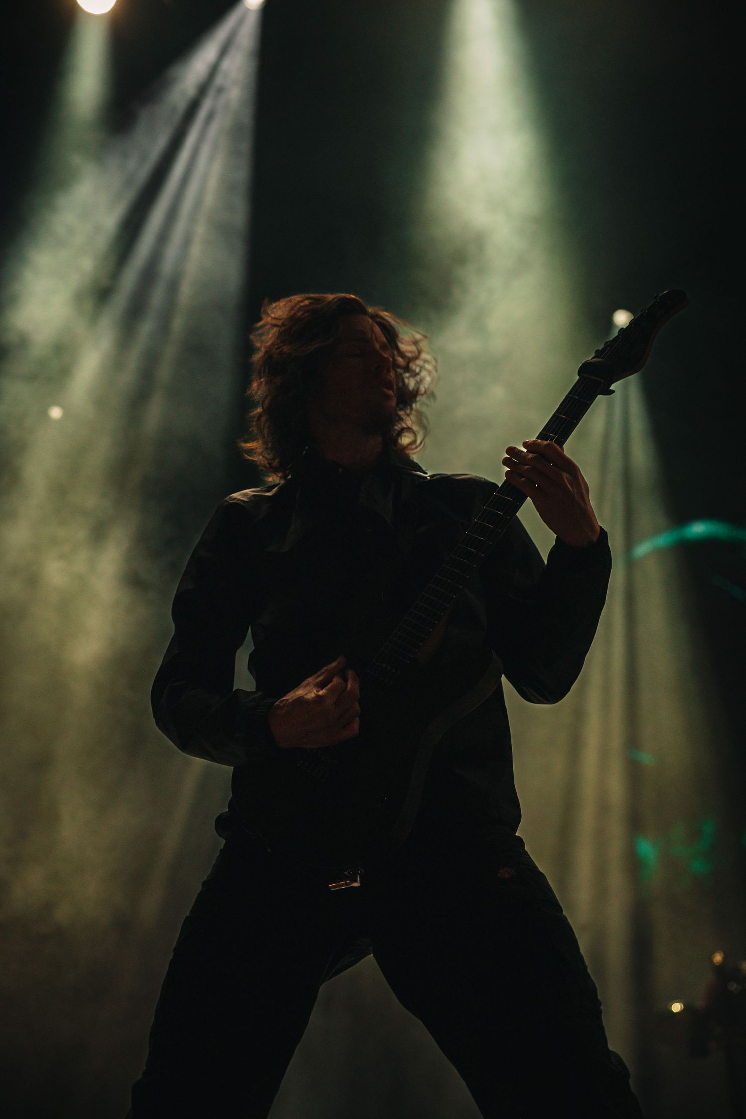 A rock guitarist performs on a smoky stage under dramatic green spotlights, playing an electric guitar in dark attire.