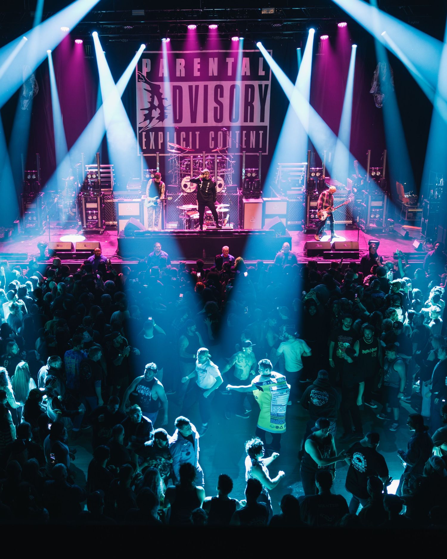 Live concert with crowd dancing under pink and blue stage lights with a Parental Advisory backdrop.