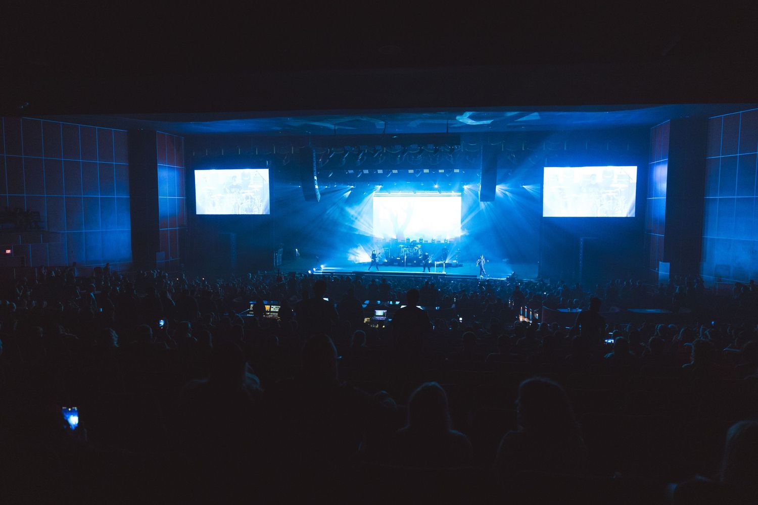 A large concert crowd bathed in blue light faces a bright illuminated stage with two large screens.