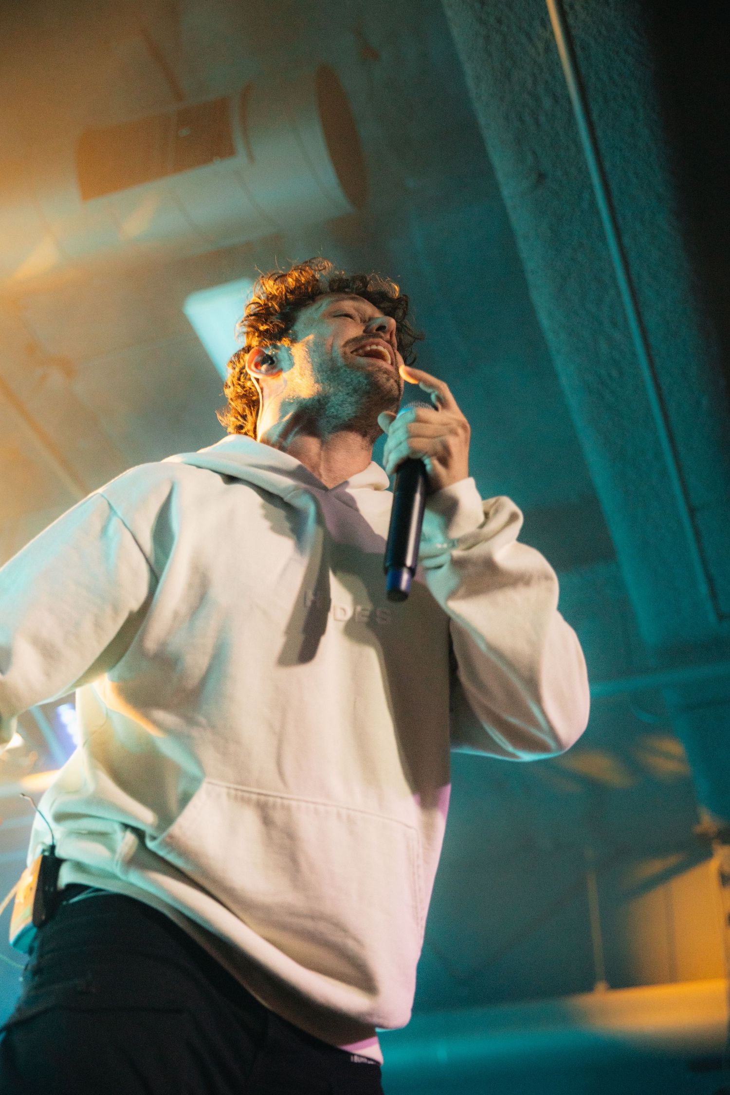 A performer in a white hoodie sings passionately on stage with dramatic turquoise lighting in the background.