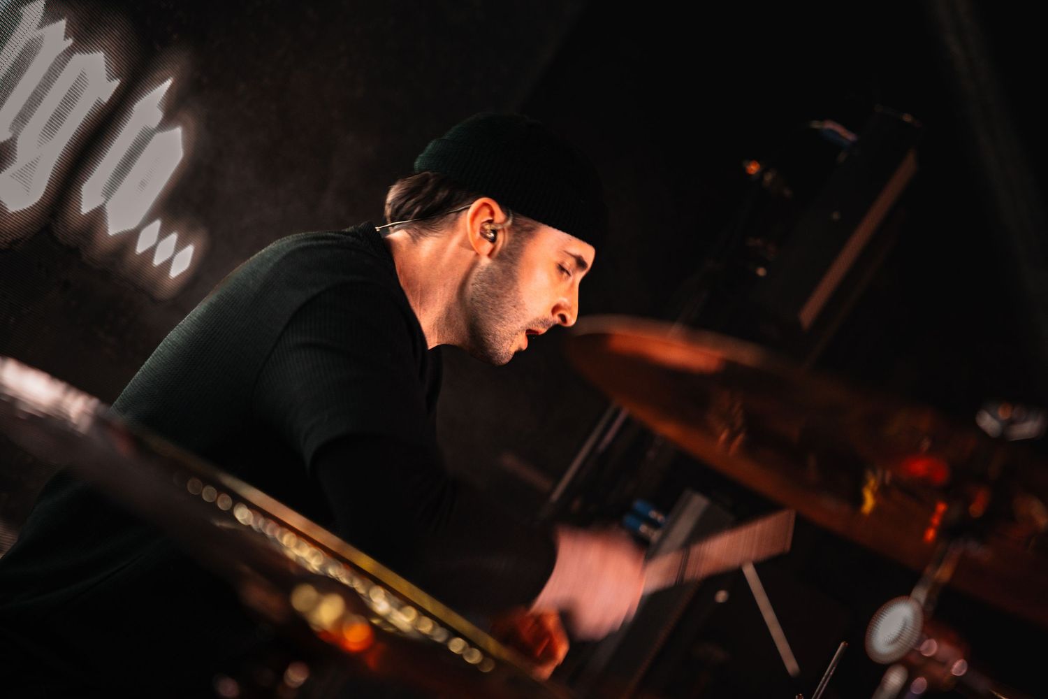 A focused male drummer wearing a black beanie performs intensely on stage under dim concert lighting.