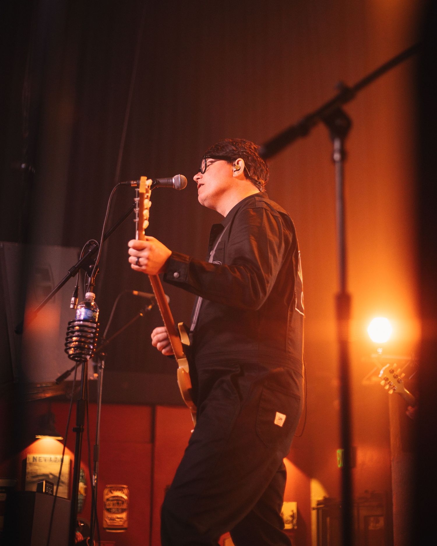 Musician in black attire performs on stage with guitar under dramatic orange lighting at a live concert venue.