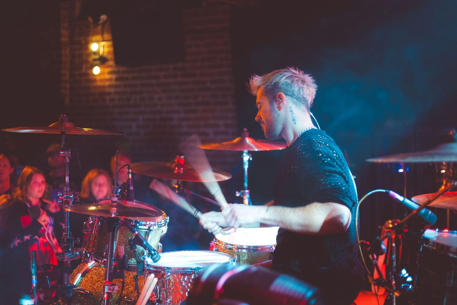 Drummer performs energetically on stage with blue lighting and brick wall backdrop.