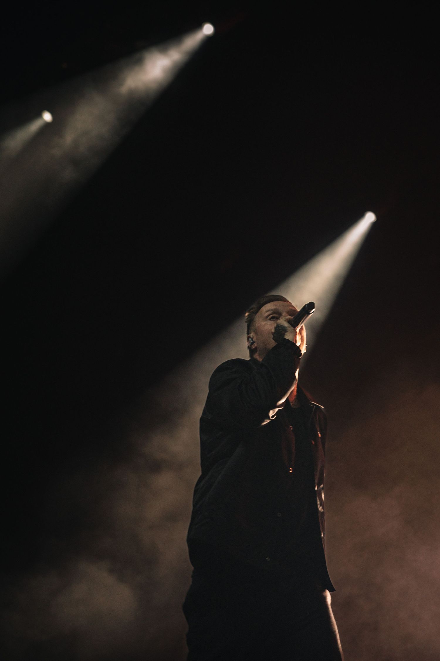 Male performer singing on dark stage with dramatic spotlights and smoke effects during live concert.
