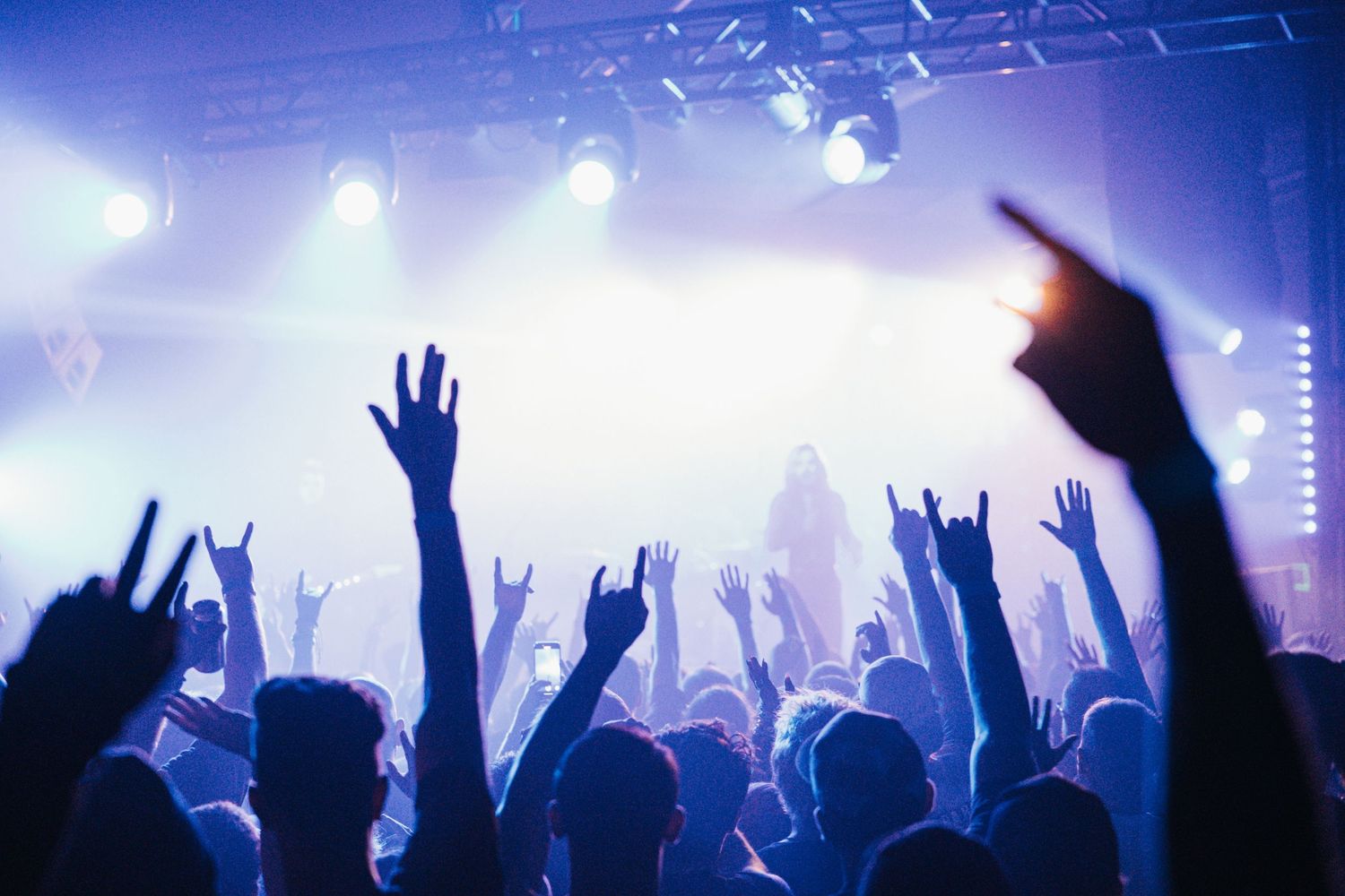 Silhouettes of raised hands at a concert with bright blue stage lighting and fog creating an atmospheric mood.