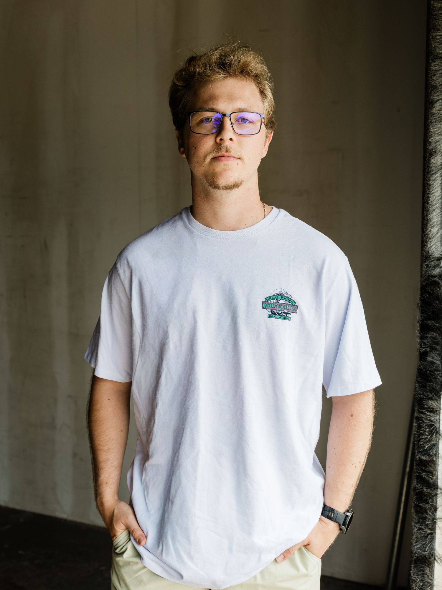 Young man with blonde hair and blue glasses wearing a white graphic t-shirt and khaki pants, standing confidently indoors.